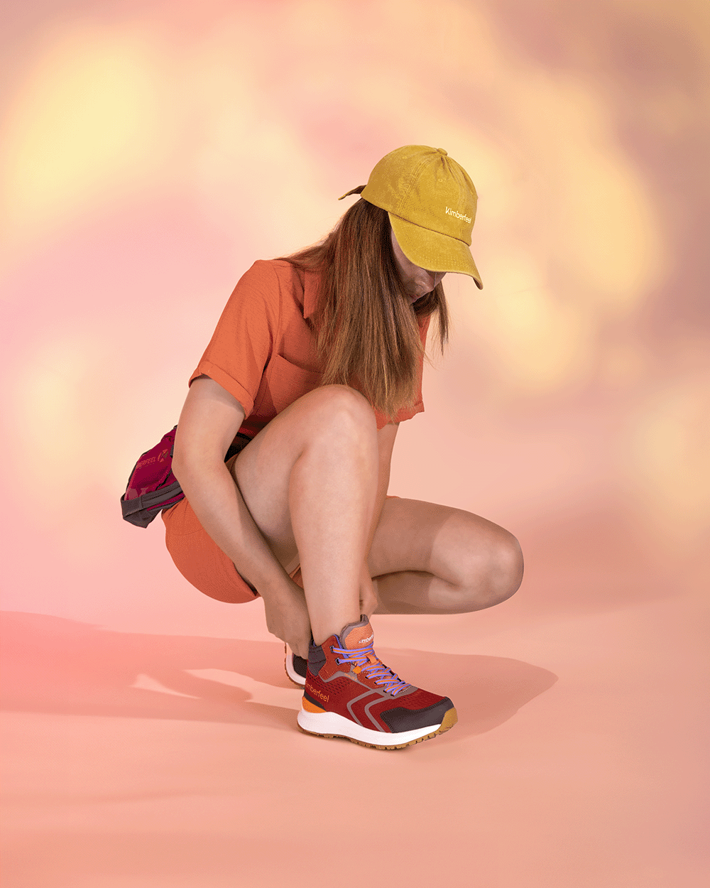 Femme accroupie en tenue orange et casquette jaune, portant les chaussures de randonnée TARAGHA rouges de Kimberfeel — style Gorpcore coloré sur fond studio pastel