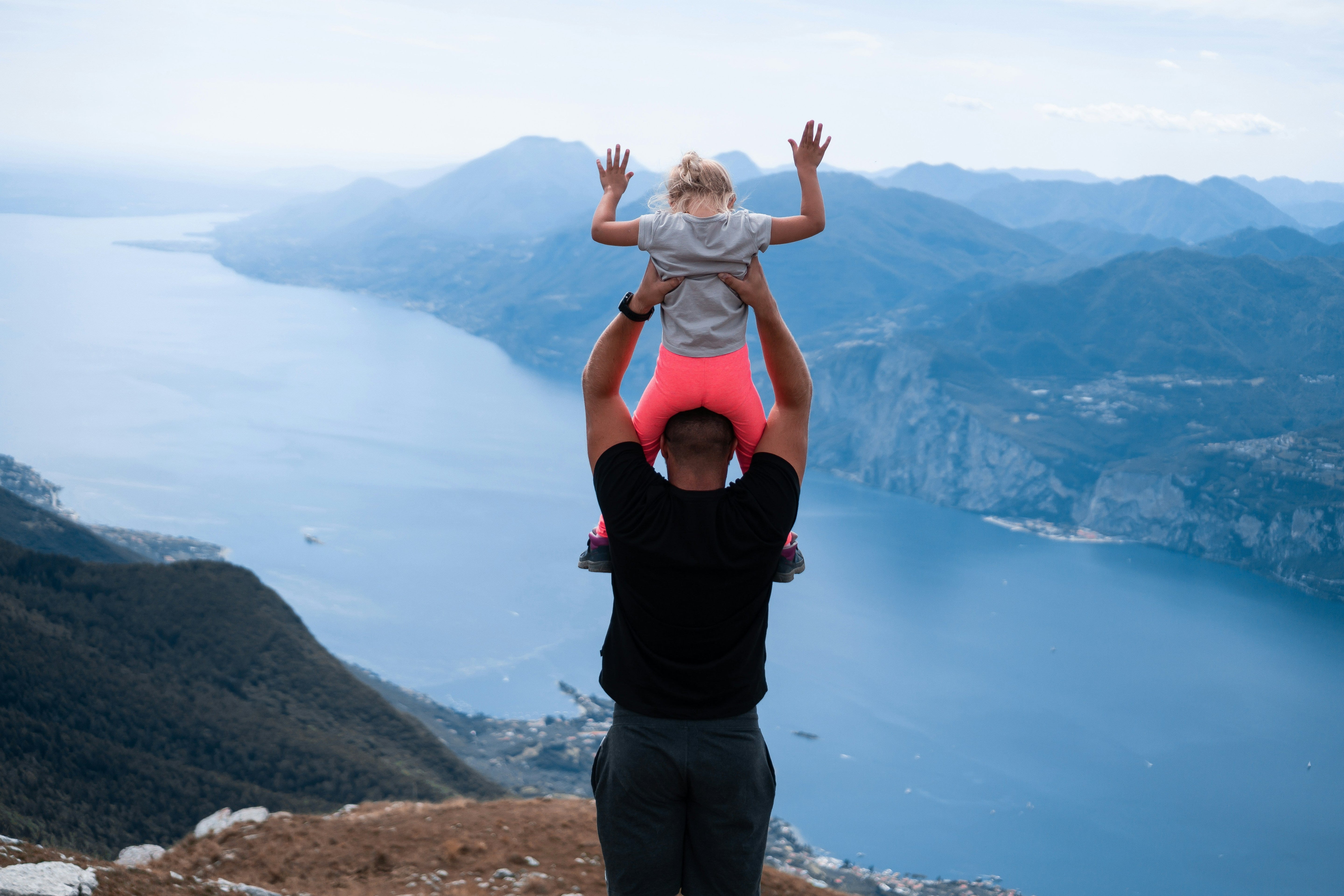 Père portant sa fille sur ses épaules en randonnée avec panorama sur lac en arrière-plan